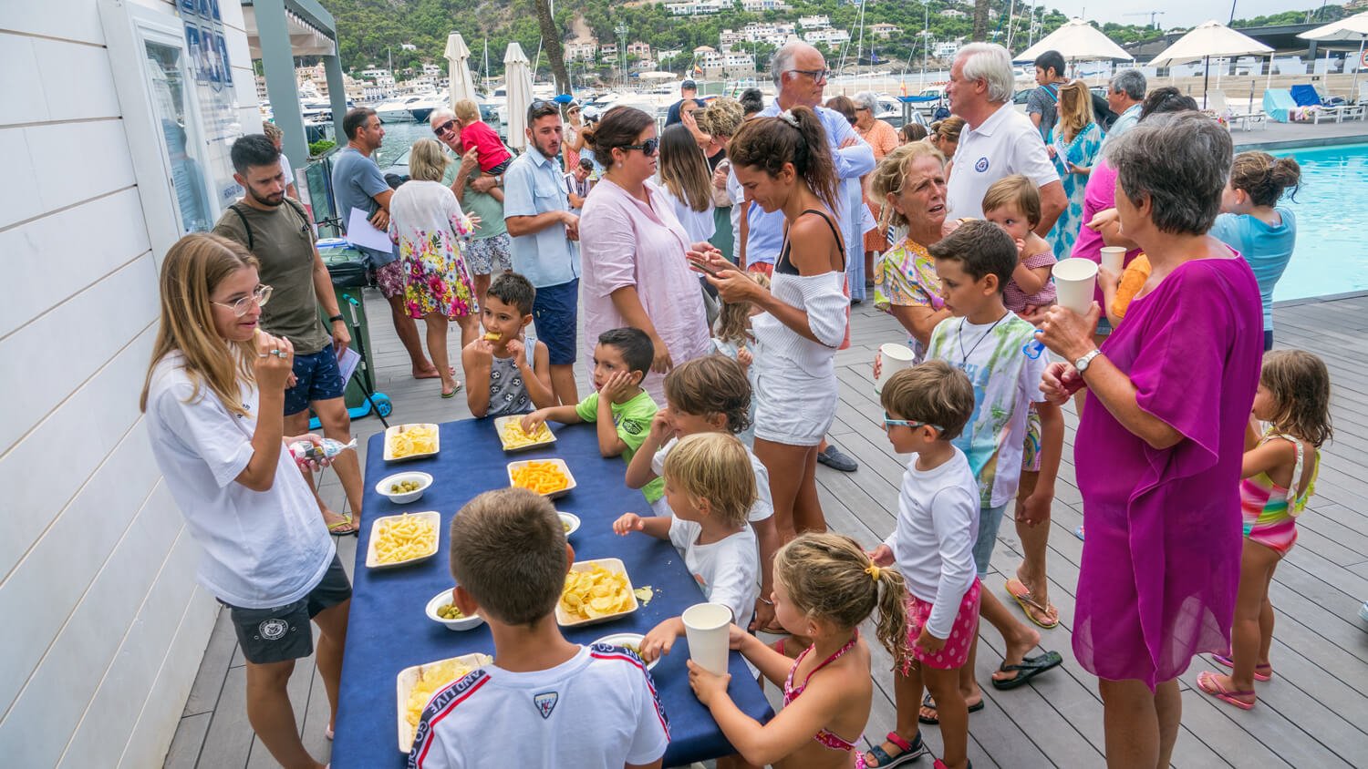 Grupo de niños y adultos en una fiesta cerca de un puerto con barcos, disfrutando de comida en una mesa cubierta con un mantel azul.