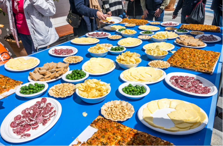 mesa con diferentes tipos de quesos, embutidos, papas, y otros aperitivos, con personas alrededor en una reunión outdoor.