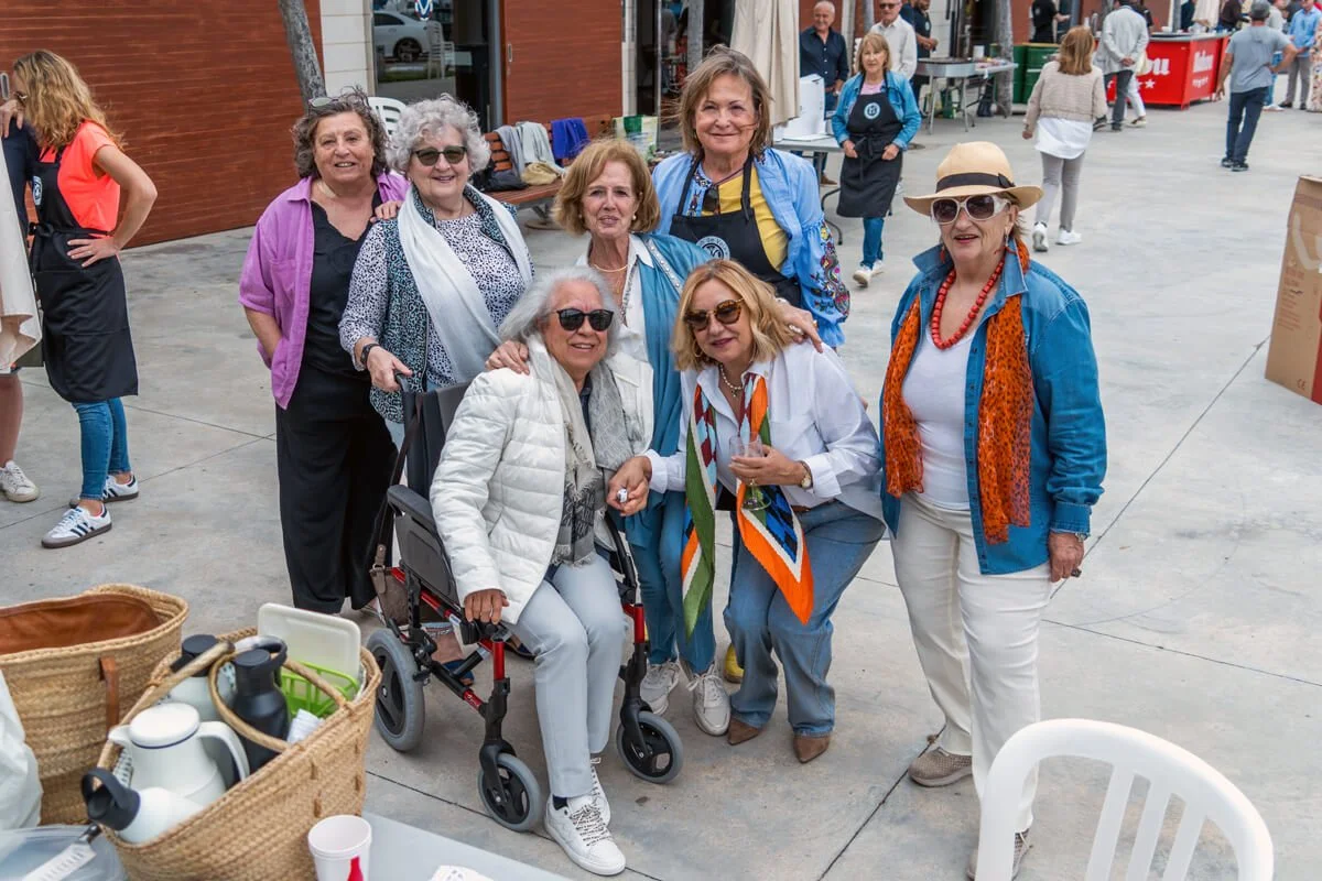 Grupo de siete mujeres adultas en un evento al aire libre sonriendo y posando juntas, con algunas usando gafas y ropa colorida, en un ambiente de mercado o feria.