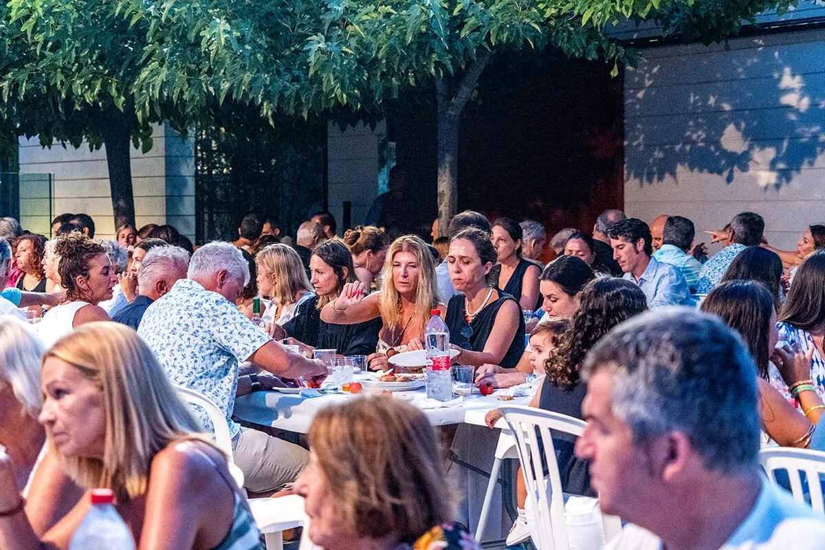 Personas sentadas en mesas durante una comida en un evento al aire libre, con árboles y una pared de fondo.
