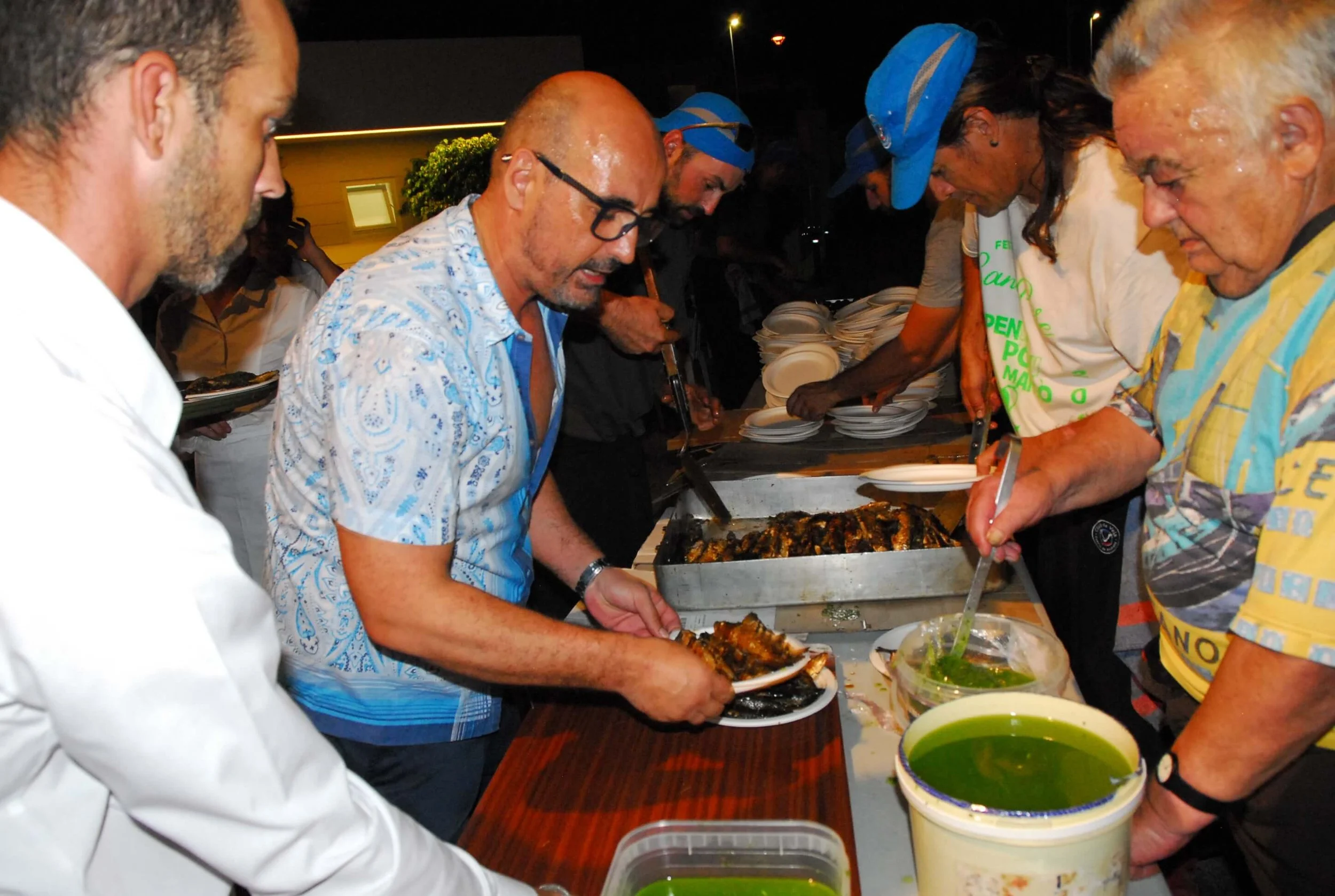Personas sirviendo comida en un evento nocturno, con platos de comida y salsas sobre la mesa.