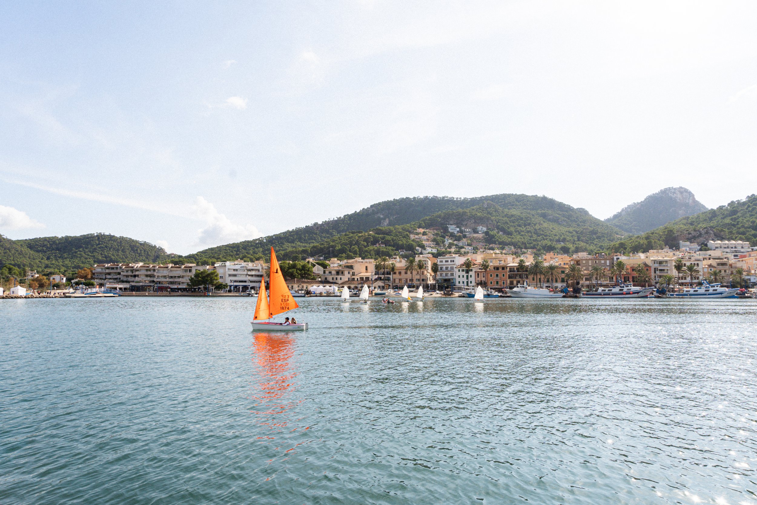 Vista de un puerto con barcos y veleros en el agua, frente a una ciudad costera con edificios y palmeras, con colinas verdes y montañas al fondo, cielo despejado.