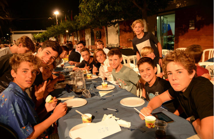 Un grupo de niños y adolescentes sentados en una mesa al aire libre durante la noche, disfrutando de una comida y refrescos en una reunión social.