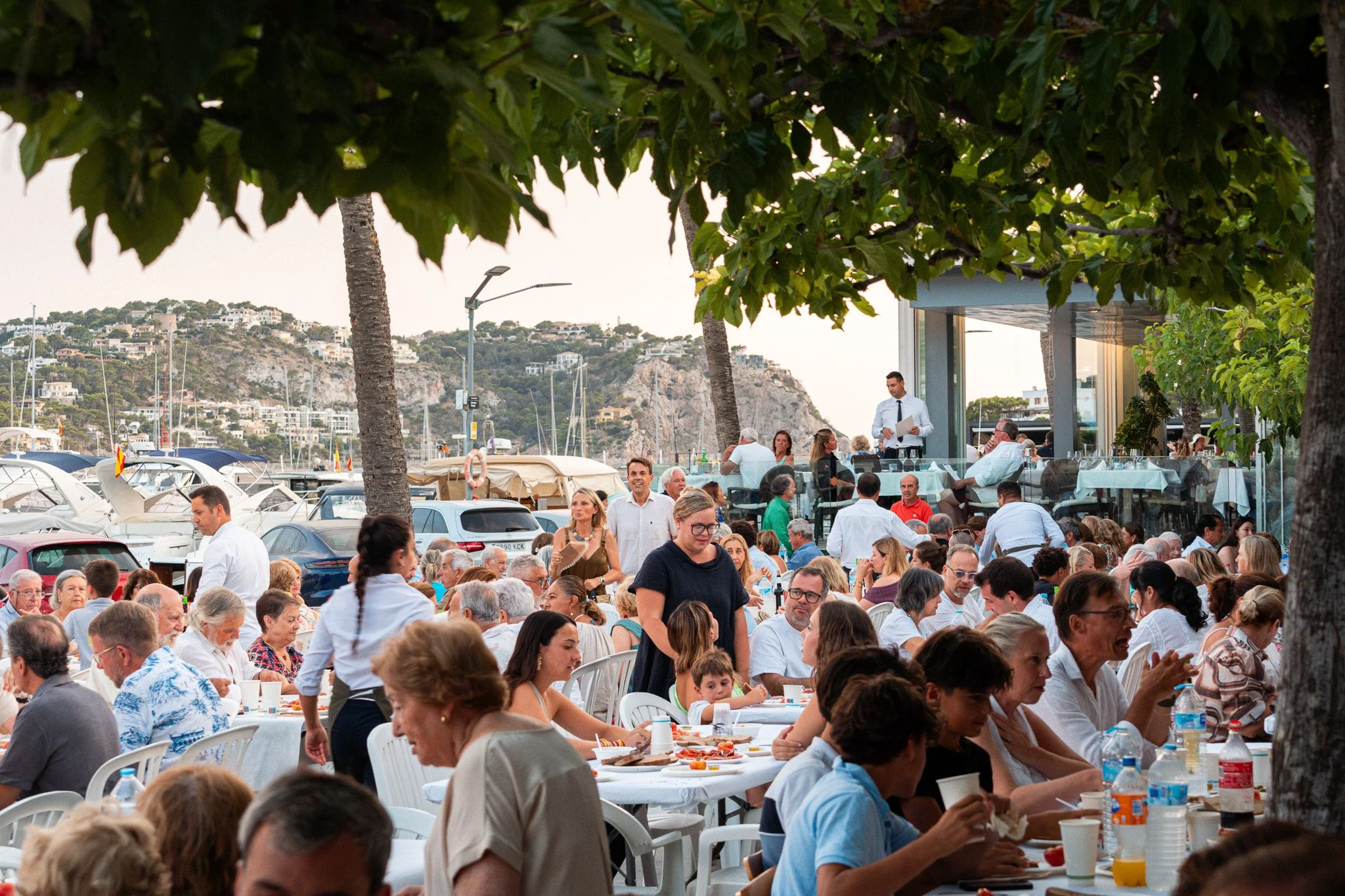 Una multitud de personas en una celebración al aire libre junto a un muelle con yates, árboles altos y colinas con casas al fondo, con mesas de comida y gente comiendo y socializando.
