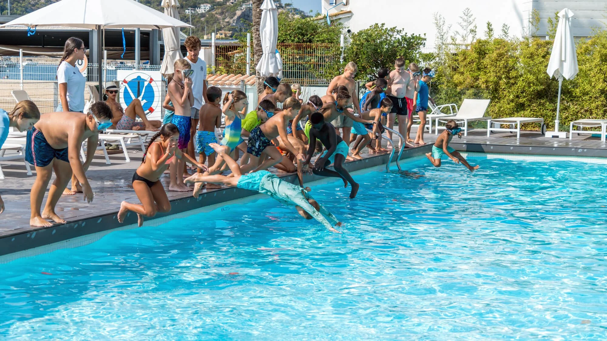 Niños en traje de baño saltando y cayendo al agua desde la piscina, mientras algunos adultos supervisan en un área aledaña al borde de la piscina bajo paraguas blancos.