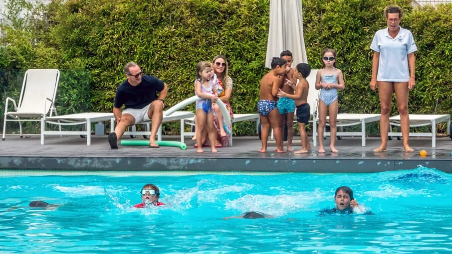 Grupo de niños y adultos en una piscina con sombra, algunos niños nadando y otros en la cuerda, mientras dos adultos supervisan desde la orilla.
