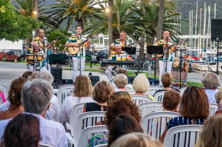 Un grupo de músicos tocando en vivo en un escenario al aire libre con palmeras, en frente de una audiencia sentada en sillas de plástico, durante la tarde.