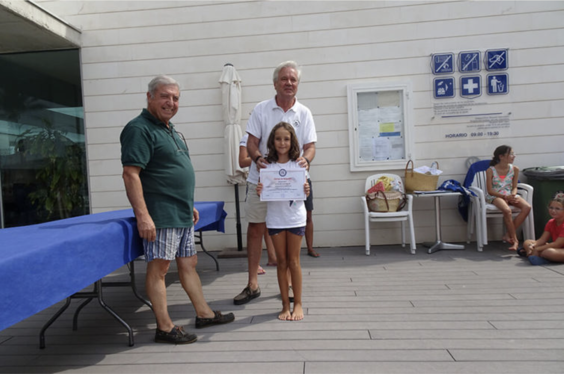 Un grupo de personas en una ceremonia al aire libre. Un niño con certificado en mano recibe un reconocimiento mientras dos adultos la acompañan y otras personas están sentadas en sillas.
