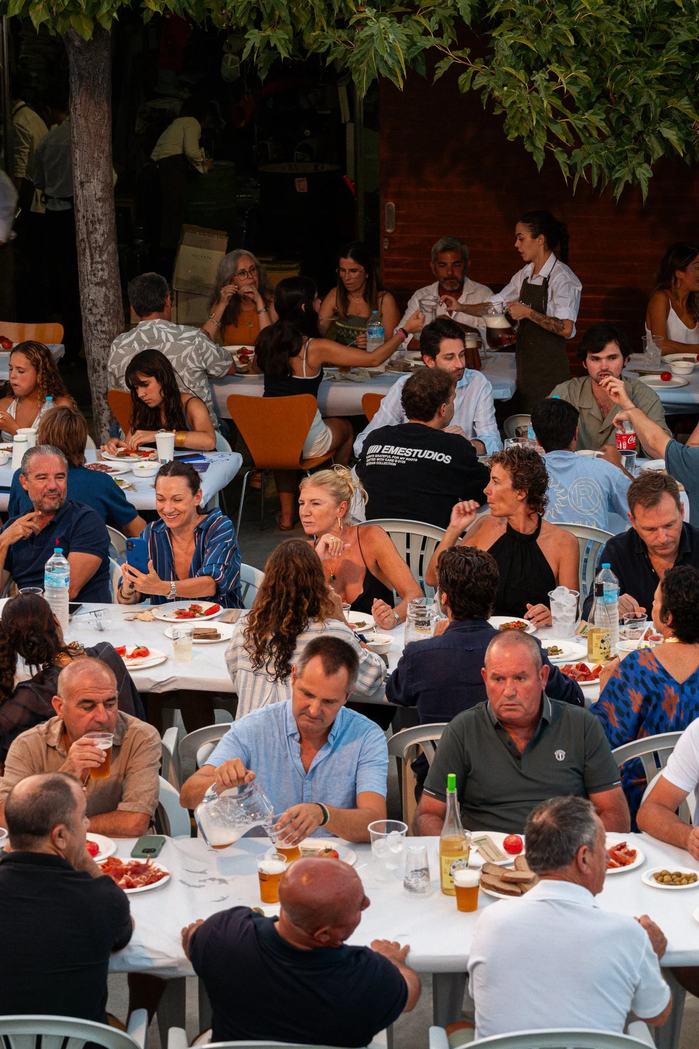 Una reunión de muchas personas sentadas en mesas durante una cena o comida al aire libre, con platos de comida y bebidas en las mesas, y una camarera sirviendo bebidas a una de las mesas.