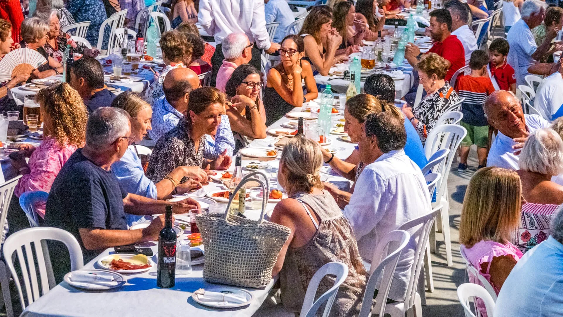 Personas sentadas en una mesa larga al aire libre, disfrutando de una comida en un evento social, con platos, botellas y vasos sobre la mesa.