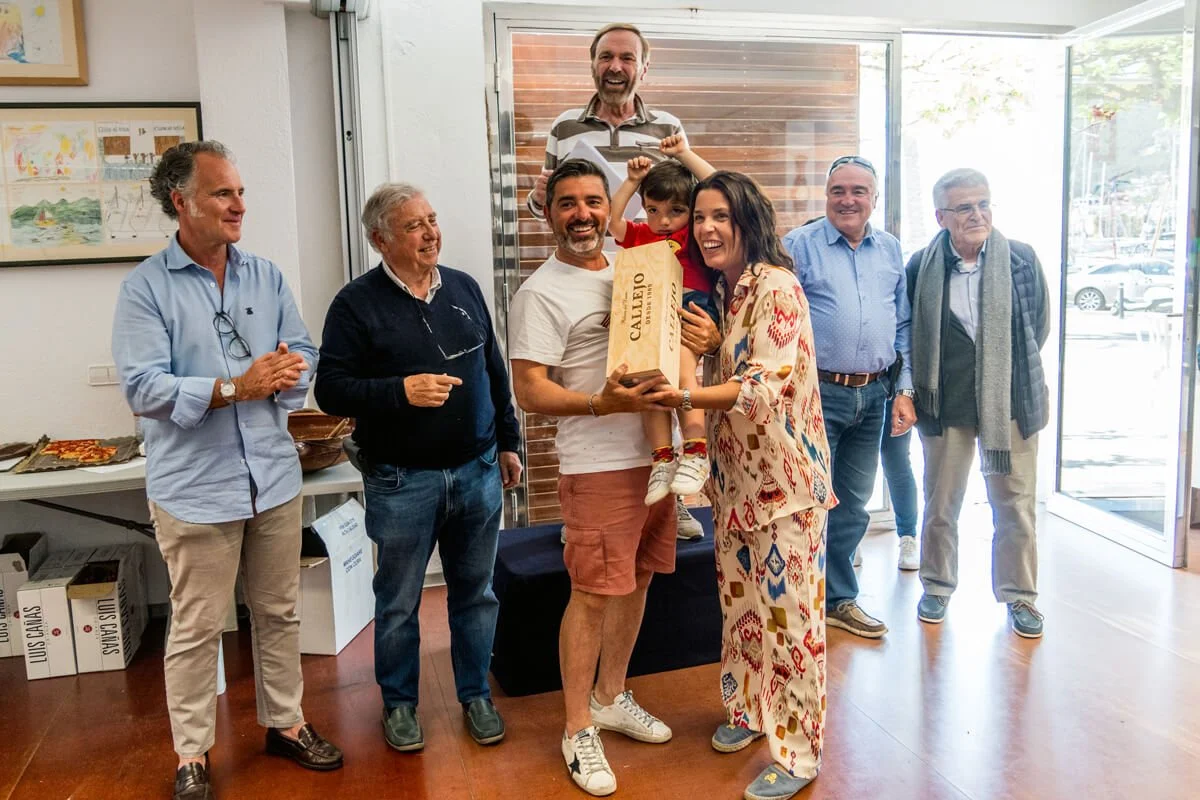 Grupo de personas en una celebración, entregándose un regalo y sonriendo, en un lugar con ventanas grandes y decoraciones en las paredes.