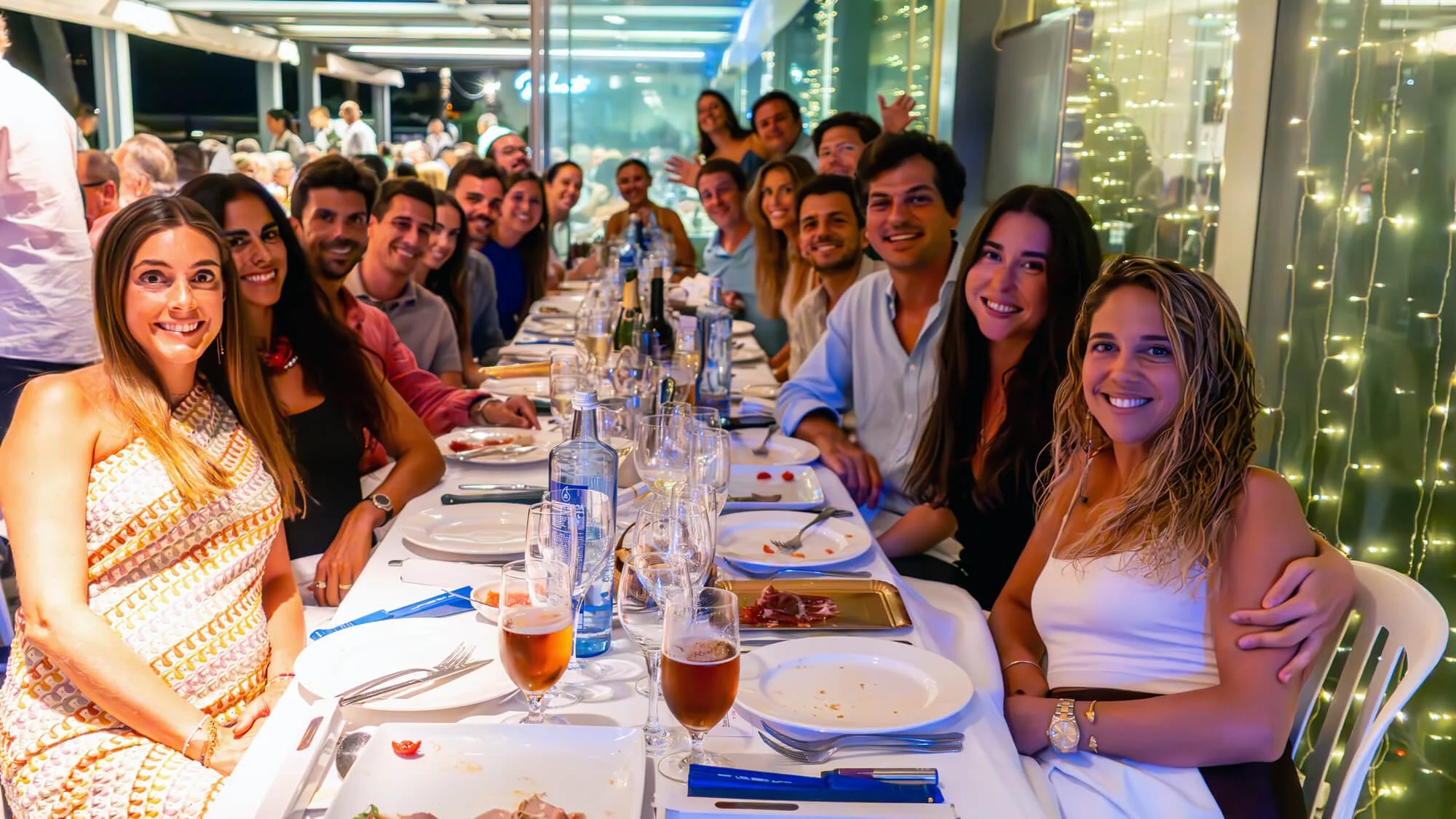 Grupo de personas sonrientes en una cena en un ambiente decorado con luces de navidad en un restaurante o terraza por la noche.