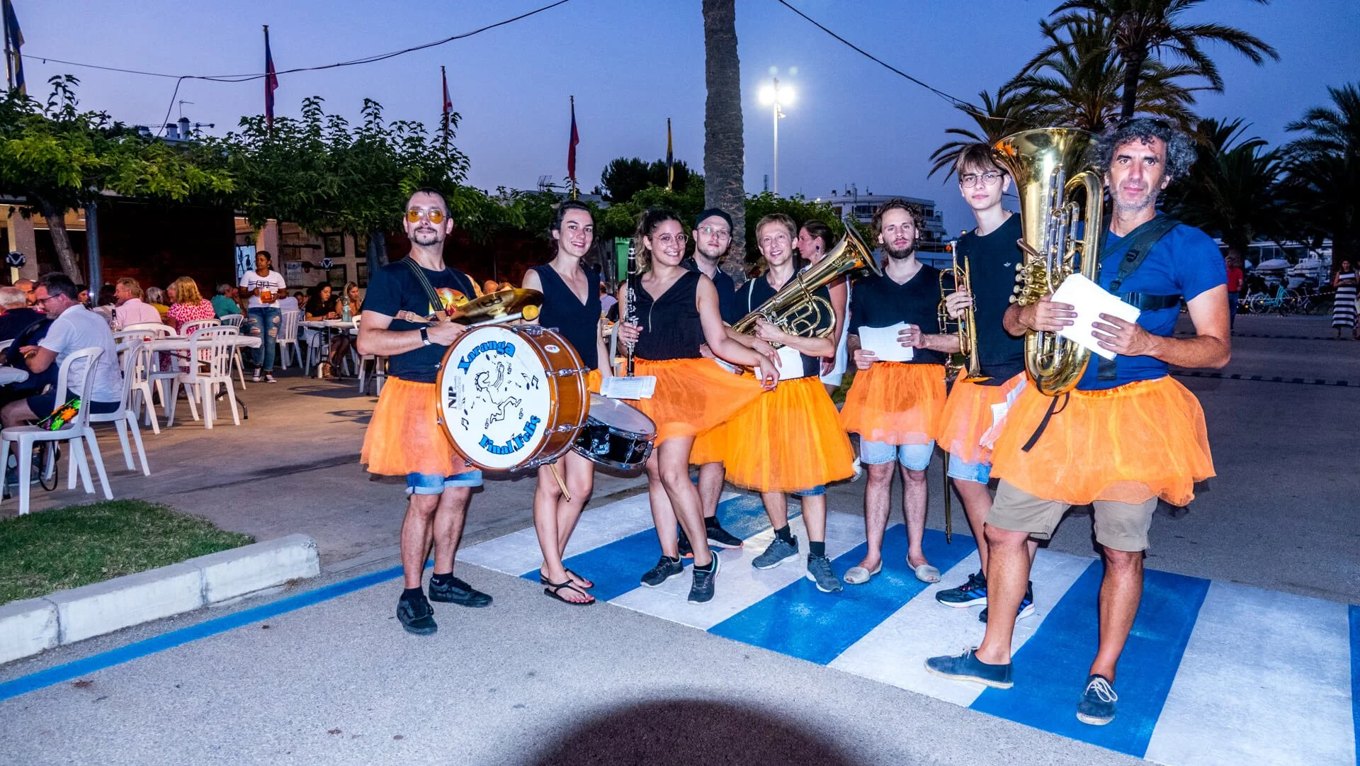 Grupo de músicos con camisetas negras, faldas de tul naranjas y diferentes instrumentos musicales en una calle durante un evento nocturno.