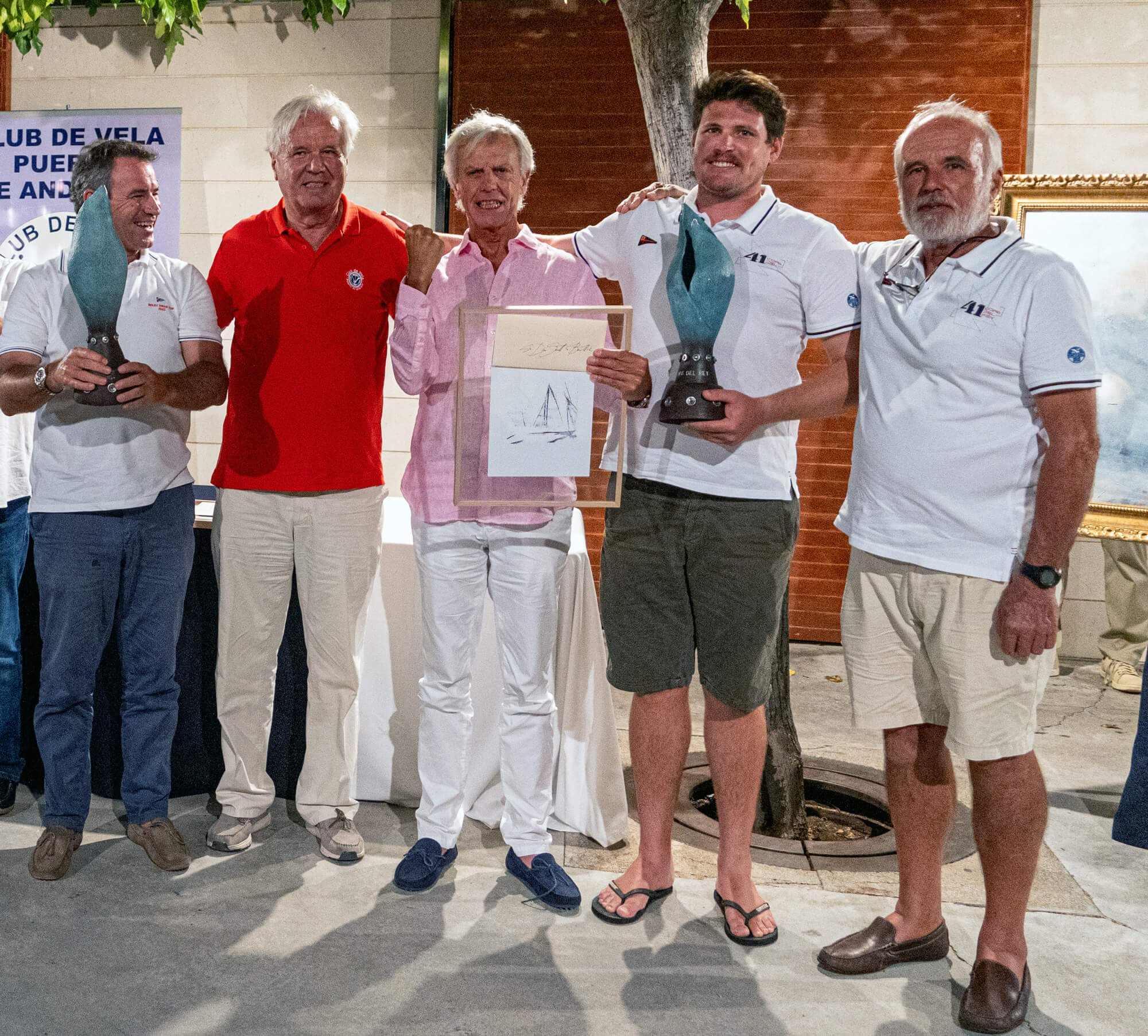 Grupo de cinco hombres en un entorno exterior, algunos sosteniendo trofeos de vela, parece ser una ceremonia o evento de entrega de premios.