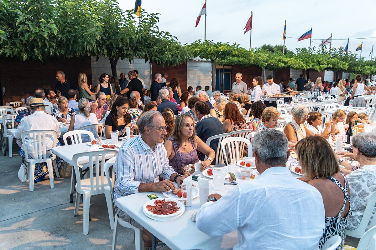 Una reunión al aire libre con muchas personas sentadas en mesas comiendo y socializando, con banderas ondeando arriba y árboles en el fondo.