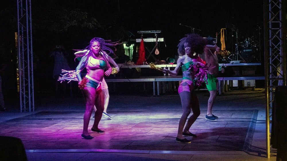 Ellas están bailando en un escenario con luces de colores, usando trajes de baño y sosteniendo pompones.