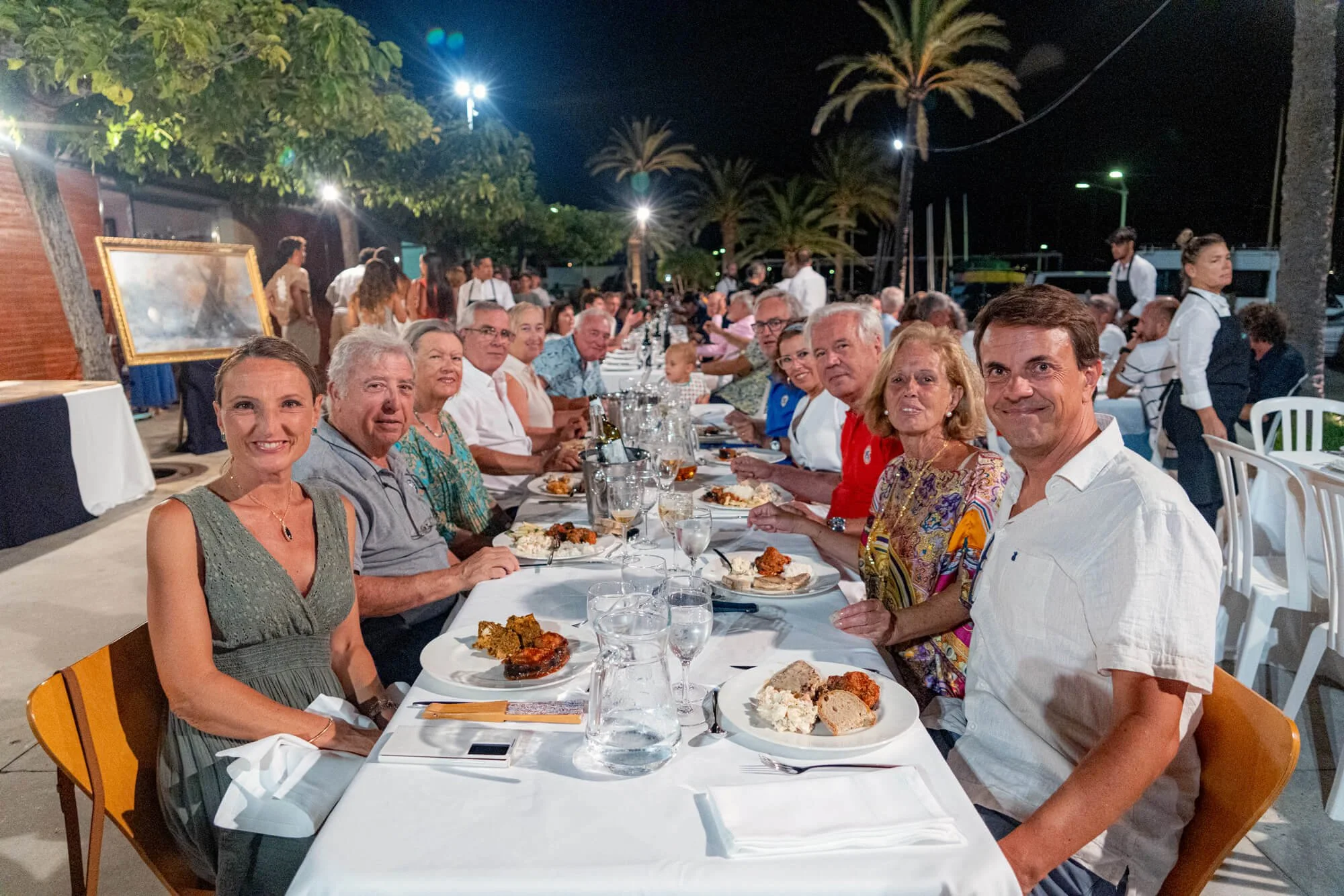 Personas sentadas en una mesa larga durante una cena nocturna al aire libre, con comida en platos, en un ambiente con palmeras y luces. Algunos comensales están sonriendo y otros parecen conversar o mirar la cámara.
