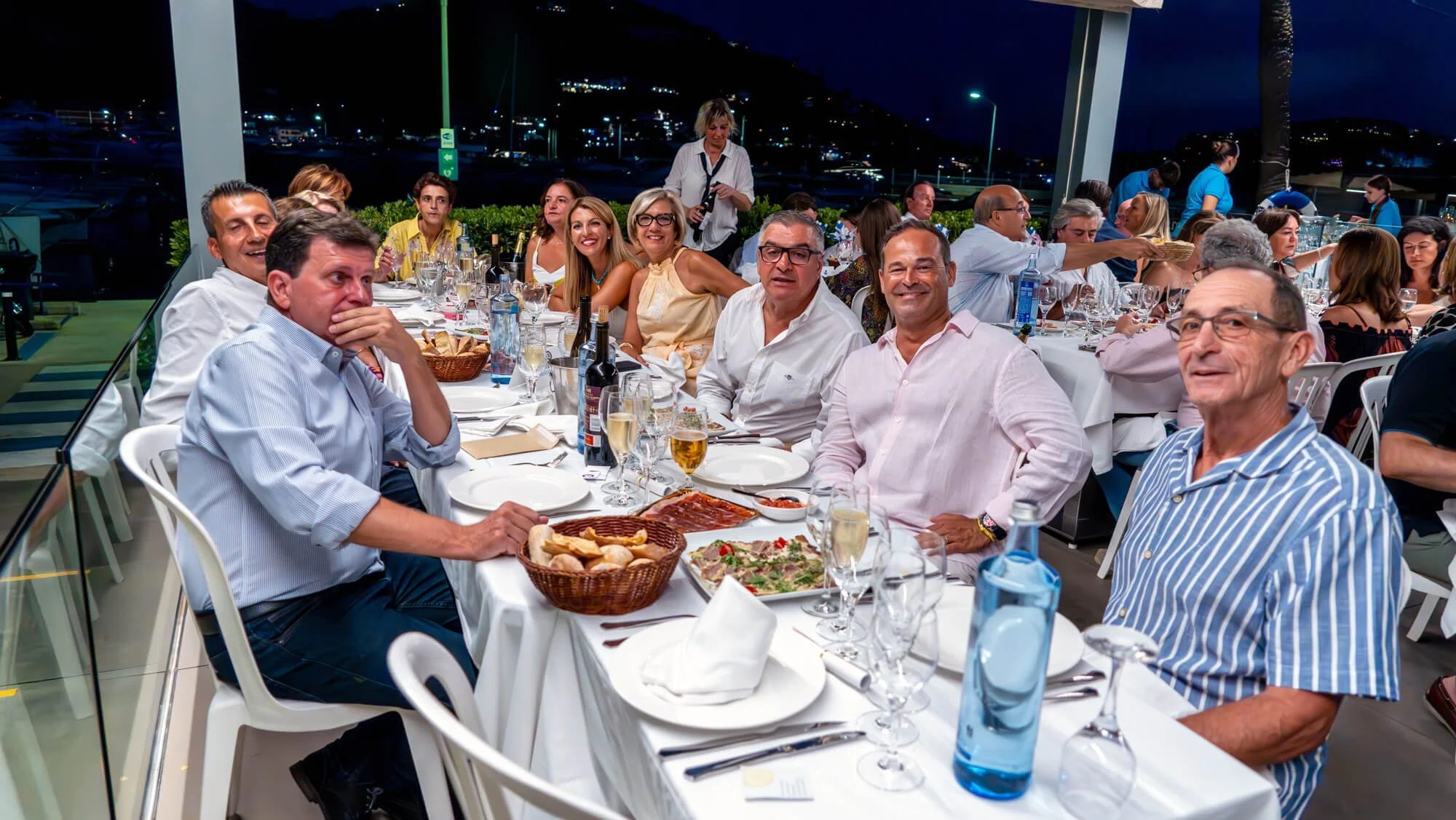 Personas sentadas en una cena al aire libre por la noche en una terraza, con platos de comida, copas de vino, y bebidas en la mesa, algunas sonriendo y otras conversando.