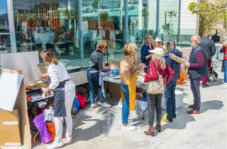 Grupo de personas en un mercado o feria al aire libre, algunos comprando y otros vendiendo productos en una mesa
