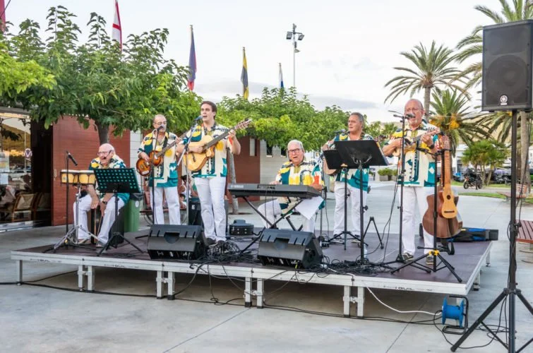 Grupo de músicos tocando en un escenario al aire libre, con instrumentos como guitarras, teclado y micrófonos, en un entorno con árboles y palmeras.
