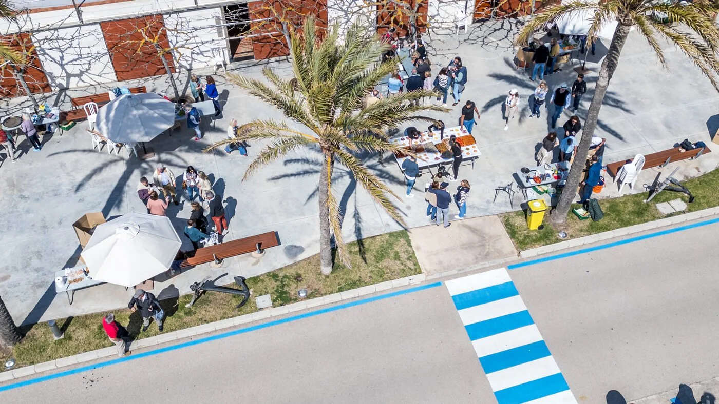 Grupo de personas en un evento al aire libre con mesas, food trucks y sombra de palmeras.