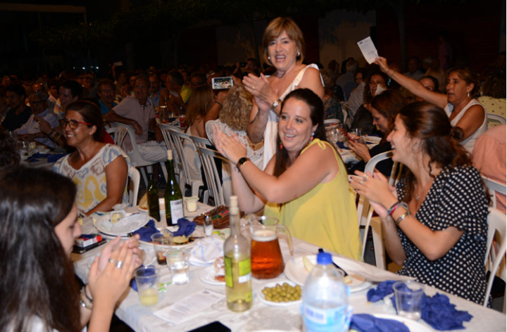 Un grupo de mujeres en una celebración disfrutando y conversando en una mesa, con bebidas y comida, en un lugar con múltiples asistentes.