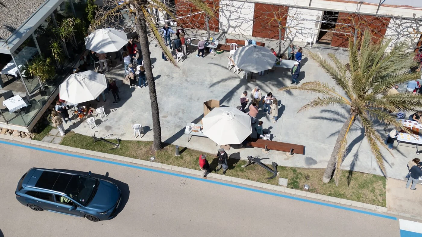Vista aérea de una terraza con mesas, sillas y sombrillas, con varias personas socializando. Hay palmeras y árboles en el área y un coche estacionado en la calle.