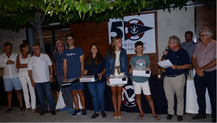 Grupo de personas en una ceremonia, algunos sosteniendo cajas y un hombre hablando con un micrófono, en un lugar decorado con un cartel que celebra el 50 aniversario de una organización.