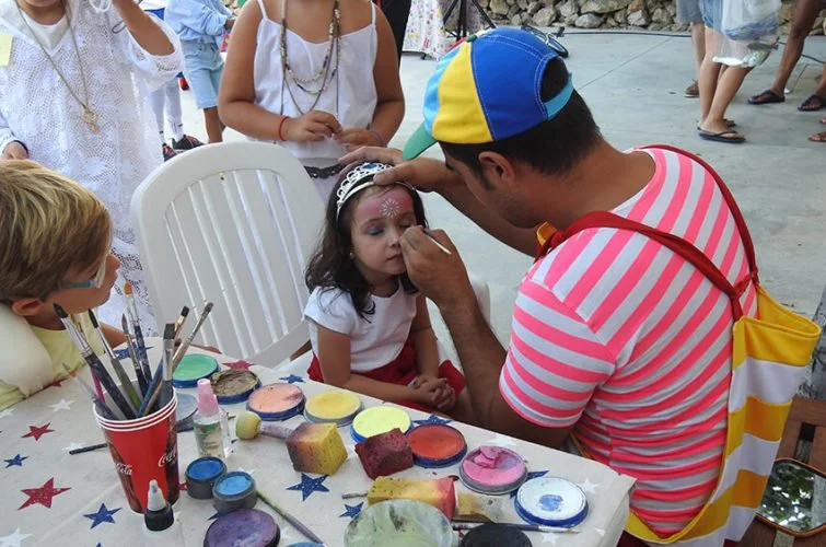 Un hombre pintando la cara de una niña en una celebración infantil al aire libre, con otros niños y adultos cercanos y coloridos objetos de pintura en la mesa.
