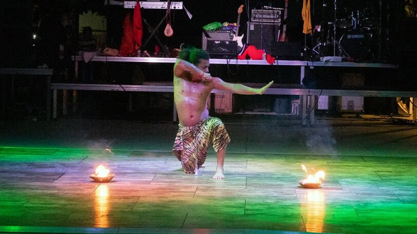 Un hombre sin camisa vestido con pantalones de tigre realiza una danza o acto en el centro del escenario, rodeado de fuego en llamas sobre el suelo, con un fondo de equipo de sonido y instrumentos musicales.