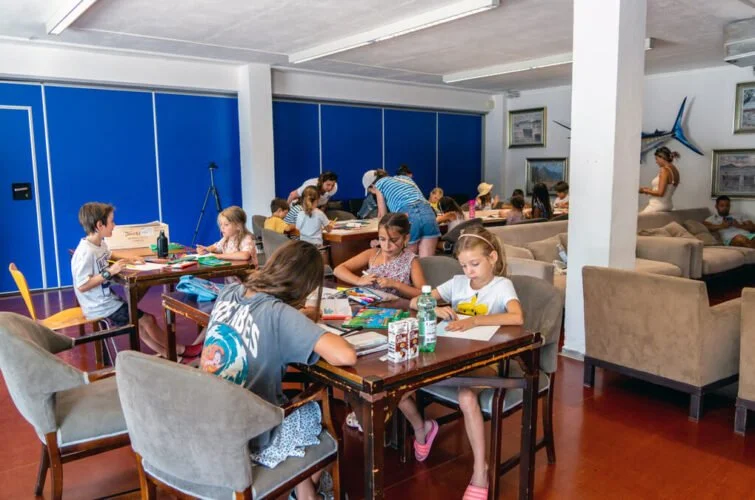 Niños y adolescentes en una sala de clases o taller, sentados en mesas, realizando actividades manuales y educativas, con algunos adultos supervisando.