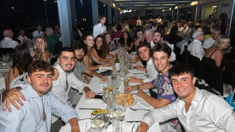 Grupo de personas sentadas en una mesa larga con comida y bebidas en un restaurante o evento social.