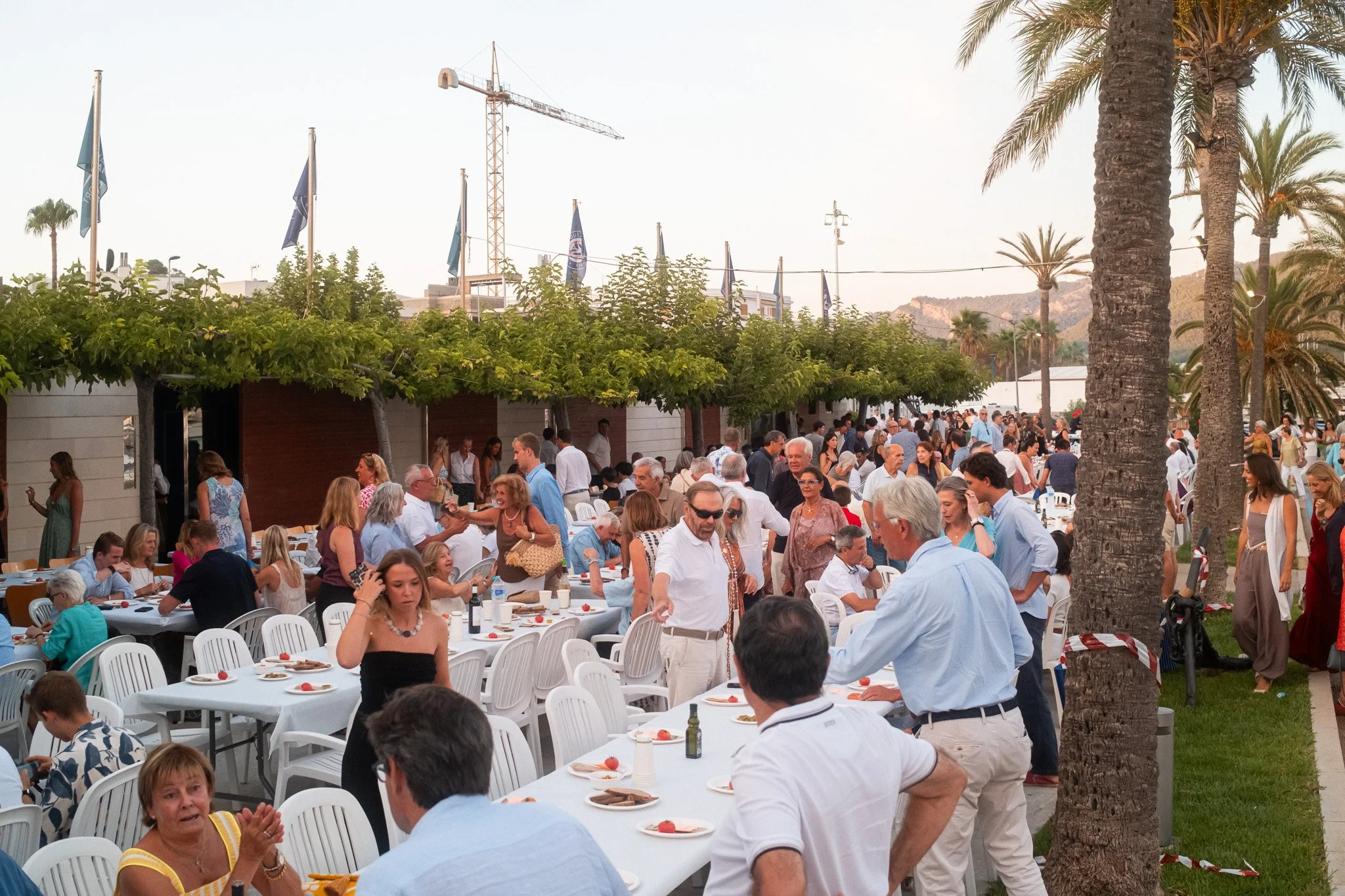 Una reunión al aire libre con muchas personas sentadas y de pie, participando en una comida o evento social, con mesas decoradas, palmeras y un cielo despejado al fondo.