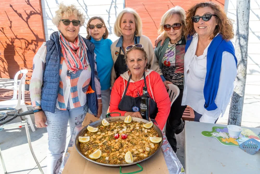 Un grupo de seis mujeres mayores sonriendo y posando cerca de una paella en una mesa al aire libre.