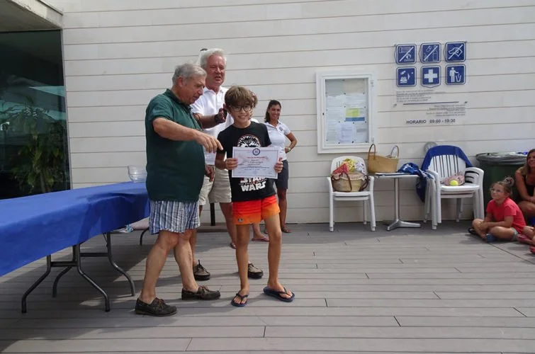 Niño joven recibiendo un certificado en una ceremonia en un espacio al aire libre, acompañado por adultos y otros niños.