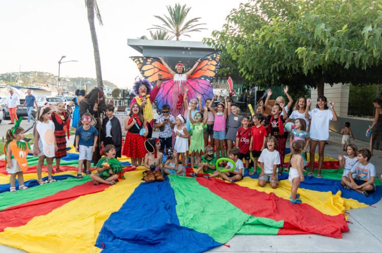 Grupo de niños y adultos en una fiesta infantil al aire libre, con un payaso y una persona disfrazada de mariposa gigante en el centro, rodeados de globos y decoraciones coloridas, en un entorno con palmeras y árboles.