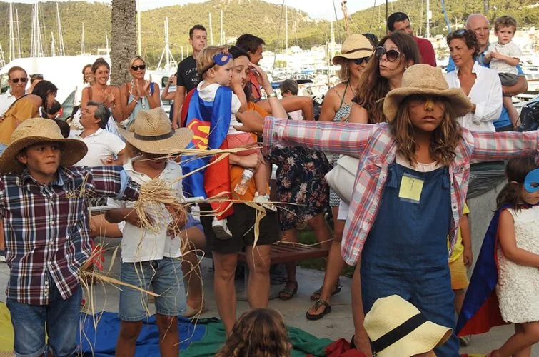 Niños disfrazados de personajes de superhéroes participando en una actividad de marionetas al aire libre, rodeados de adultos y otros niños en un puerto con yates y naturaleza de fondo.