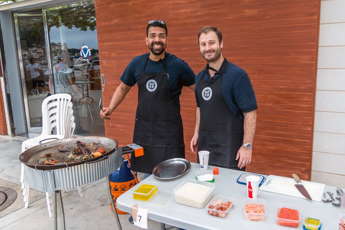 Dos hombres cocinando mariscos a la parrilla en una mesa con ingredientes y utensilios, en un espacio al aire libre frente a un edificio con paredes de madera.