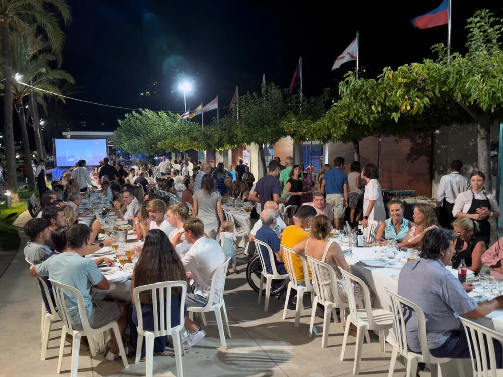 Cena al aire libre con muchas personas comiendo y socializando en una fiesta o evento nocturno, con banderas en el fondo y un gran proyector