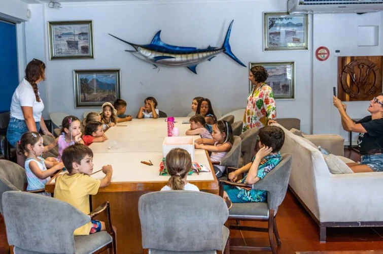 Un grupo de niños y dos adultos en una sala con decoraciones de tema marino, incluyendo un pez vela colonando la pared y varias pinturas de paisajes. Los niños están sentados alrededor de una mesa, y una mujer está de pie a la izquierda, mientras otr
