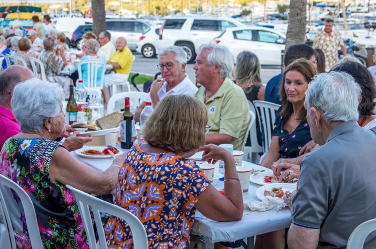 Grupo de personas mayores cenando en una reunión al aire libre con comida y bebidas.