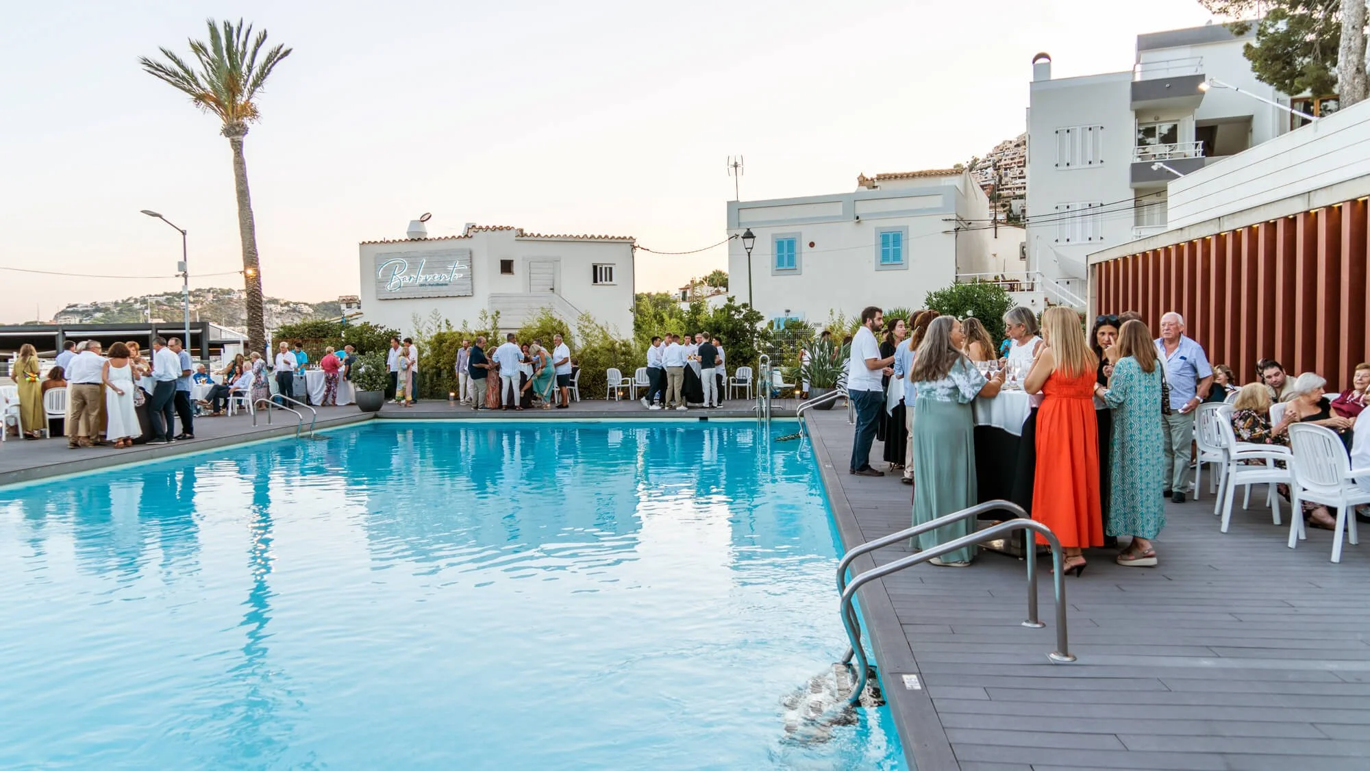 Celebración junto a una piscina con muchas personas conversando y disfrutando en una terraza con mesas y sillas, entorno urbano con edificios blancos y palmeras.