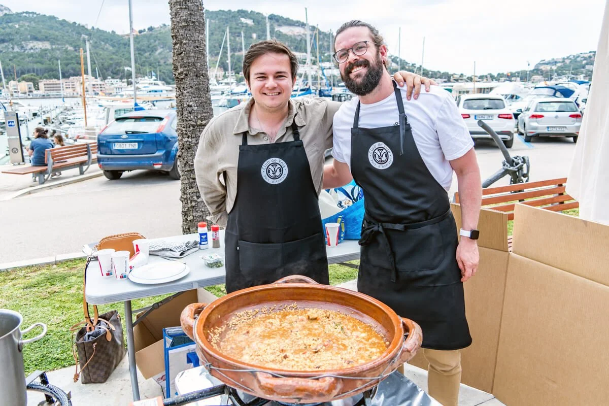 Dos hombres con delantales de cocina en una feria junto al puerto, sonriendo y mostrando una olla con paella.
