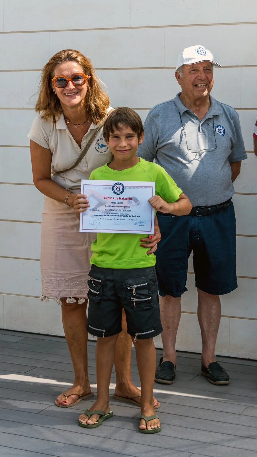 Un niño sostiene un certificado de participación en un curso de natación, acompañado de dos adultos, en un evento al aire libre.