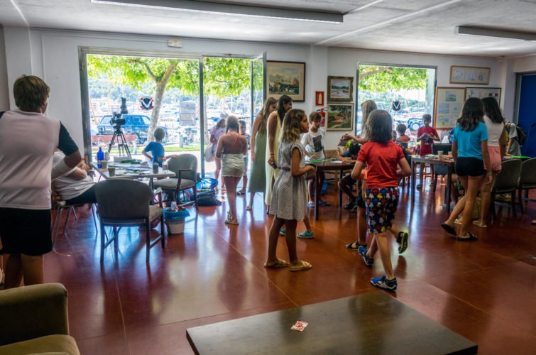 Niños y adultos en una sala de actividades, algunos esperan y otros participan en un taller, con ventanas que muestran un área exterior con árboles y un parque.