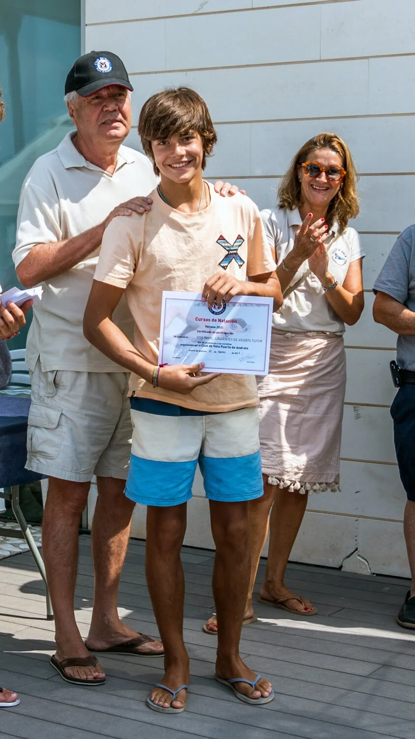 Un joven recibe un certificado de natación en una ceremonia al aire libre, rodeado de adultos que lo felicitan.