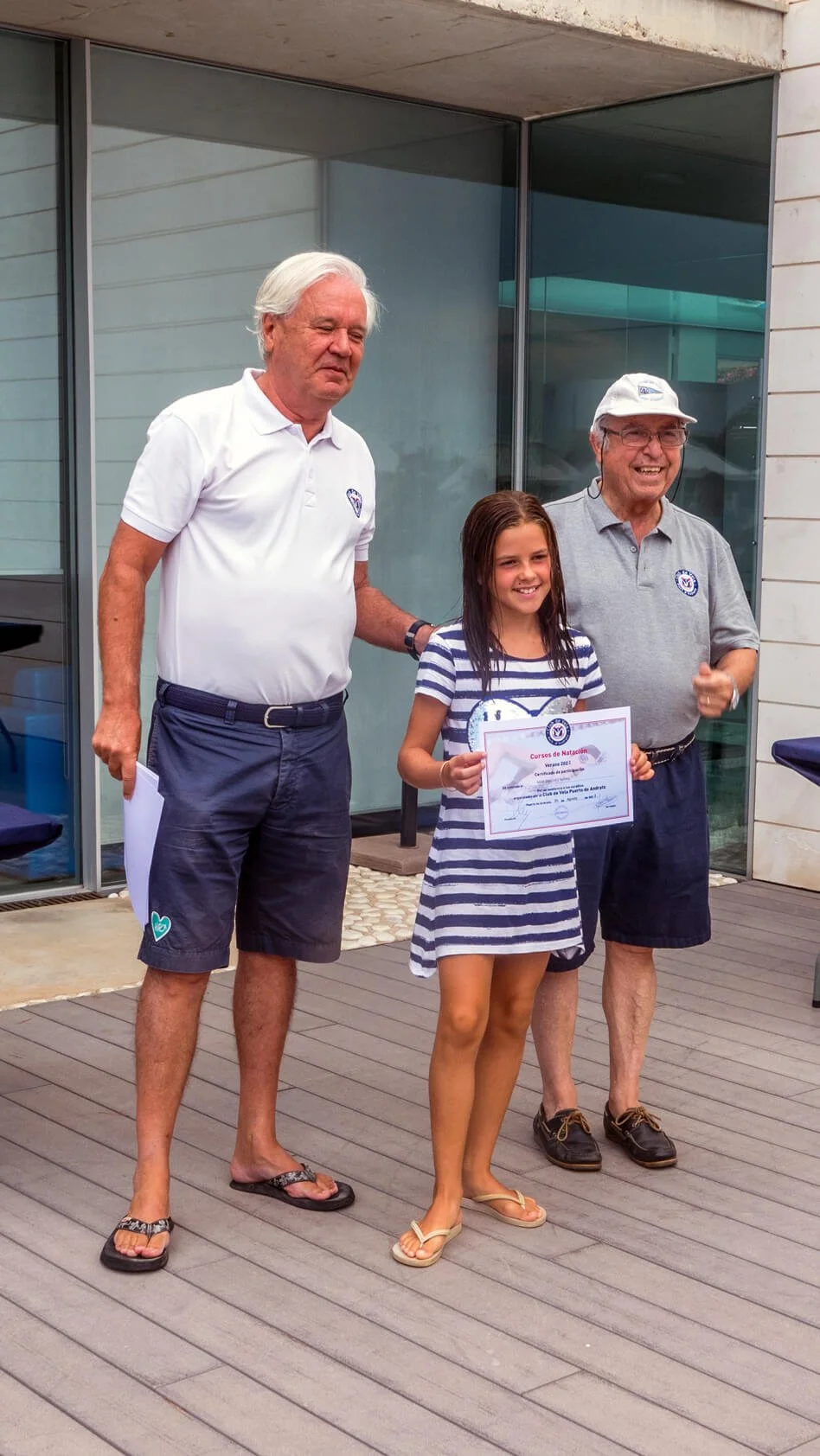 Niña sonriendo recibiendo un certificado en una ceremonia al aire libre, acompañada por dos adultos, en un entorno moderno con paredes de vidrio y madera.