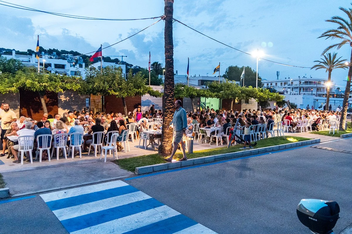 Cena al aire libre con muchas mesas y sillas blancas llenas de personas disfrutando una comida en la noche, con palmeras y edificios en el fondo.