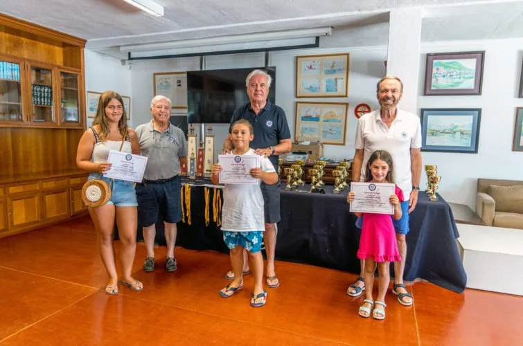 Cuatro niños y dos adultos en una ceremonia de premiación, sosteniendo certificados, en una sala con premios en una mesa y cuadros en las paredes.