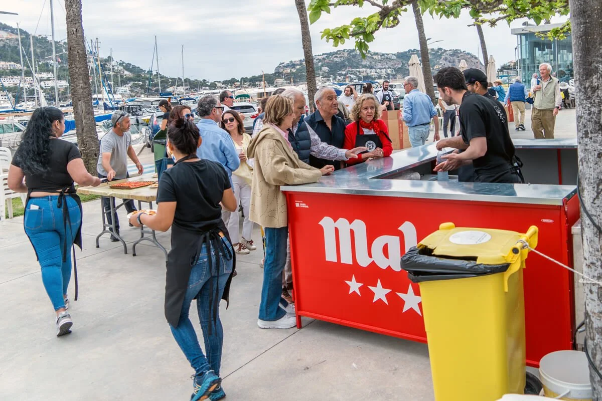 Personas comprando comida en un puesto en un muelle con barcos y colinas de fondo.
